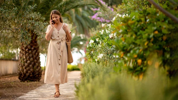 Neşeli beyaz kadın, çalılar ve palmiyeler arasında resimli asfalt yolda yürüyor ve akıllı telefondan konuşuyor. Yaz elbisesi giymiş mutlu bir kadın arkadaşına inanılmaz bir yolculuktan bahsediyor..