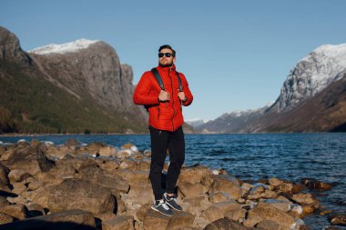 Norveç 'teki fiyortta taşların üzerinde tek başına duran kırmızı ceketli sakallı adam. Kuzeye tatile giden memnun bir adam. Seyahat yaşam tarzı duygu tatilleri, İskandinav doğa konsepti 