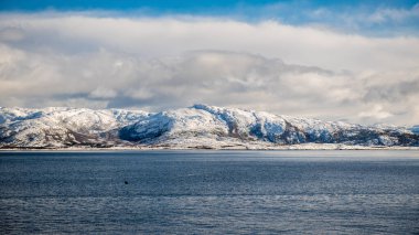 Sakin bir denizin altında bulutlu bir gökyüzünün altında karla kaplı dağlar. Lofoten Adaları, Norveç