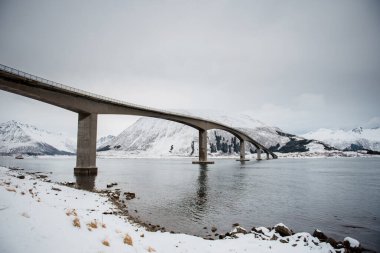 Kıvrımlı bir köprü karlı bir nehrin üzerinde uzanır ve arkasında dağlar vardır..