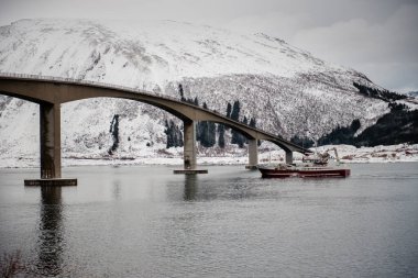 Karlı bir dağ manzarasında, beton bir köprünün altından kırmızı bir tekne geçiyor..
