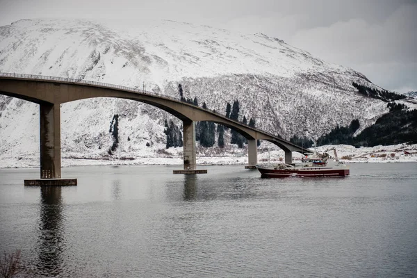 Karlı bir dağ manzarasında, beton bir köprünün altından kırmızı bir tekne geçiyor..