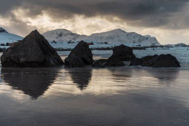 Karla kaplı dağlar ve kayalar bulutlu bir gökyüzünün altında sakin sularda yansıyor..
