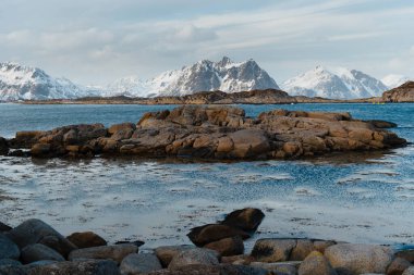 Arkasında karla kaplı dağlar olan kayalık kıyı şeridi. Lofoten Adaları, Norveç