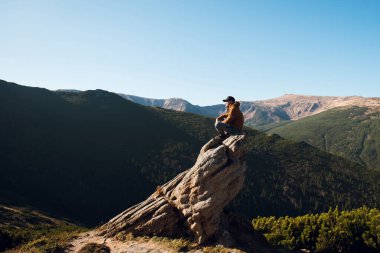 Dağ manzarasına bakan bir kayanın üzerinde oturan kişi.
