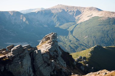 Engebeli tepeleri ve açık mavi gökyüzünün altında yeşil vadileri olan kayalık dağ manzarası.