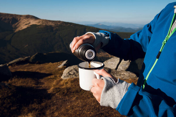 Person pouring hot beverage from thermos into mug on mountain.