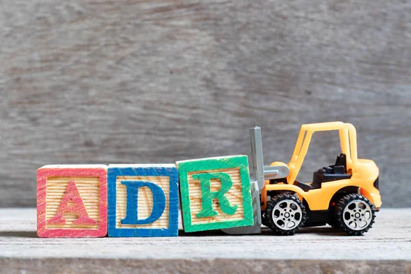 Spielzeuggabelstapler halten Buchstabenblock r, um das Wort adr (Abkürzung für unerwünschte Medikamentenreaktion) auf Holzgrund zu vervollständigen — Stockfoto