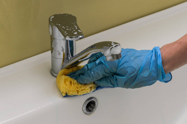 A woman in gloves washes the plumber and a tap with a rag in the bathroom with household chemicals