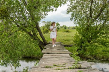 Genç ve çekici bir kız, elinde papatya buketiyle küçük bir nehrin üzerindeki eski ahşap köprüde yürüyor.