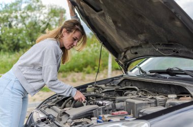 Genç ve çekici bir kız bozuk bir arabanın kaputunun yanında onu tamir etmeye çalışıyor.