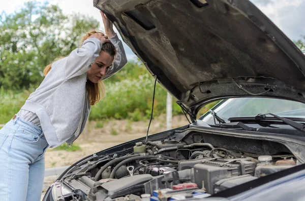 Young charming girl stands near the open hood of a broken car trying to repair it