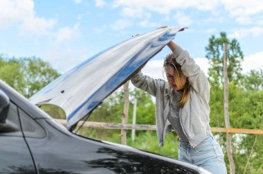 Genç ve çekici bir kız bozuk bir arabanın kaputunun yanında onu tamir etmeye çalışıyor.