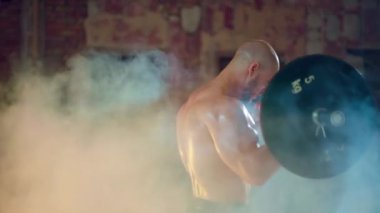 Çıplak gövdeli atletik bir adam yıkılmış bir binada duman içinde dururken bisepslerini kıvırıyor.