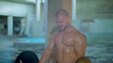 Çıplak gövdeli atletik bir adam yıkılmış bir binada duman içinde dururken bisepslerini kıvırıyor.