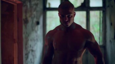 Çıplak gövdeli atletik bir adam terk edilmiş bir binada duruyor.