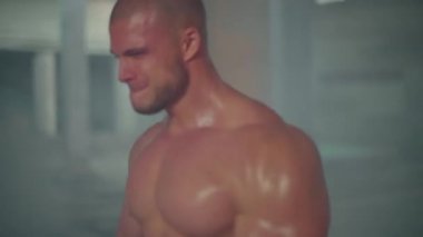 Çıplak gövdeli atletik bir adam yıkılmış bir binada duman içinde dururken bisepslerini kıvırıyor.