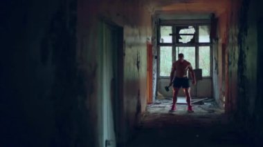 Elinde çekiçle çıplak gövdesi olan atletik bir adam yıkılmış bir binanın koridorunda duruyor.