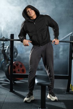 Baskı halterinin yanında kapüşonlu sweatshirt ve jimnastik kıyafeti giymiş bir adam duruyor. Formunun bir parçası olarak ağırlık kaldırmaya hazır ve odaklanmış görünüyor..
