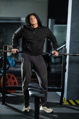 Kapüşonlu bluz giyen bir adam halterin yanında duruyor. Modern bir spor salonunda ağırlık kaldırmaya hazır..