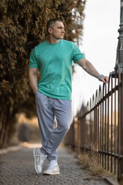 Orta yaşlı Avrupalı bir adam, turkuaz bir tişört ve parkta dekoratif bir çite yaslanmış pembe bir eşofman giyiyor. Öğleden sonra ortalık sakin..