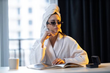 Ferahlatıcı bir duştan sonra kız mutfakta gözlük takıp rahatlıyor. Şafağın yumuşak ışınları mekanı aydınlatıyor ve sakin bir atmosfer yaratıyor..