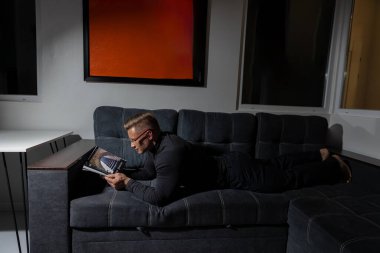 Gözlüklü atletik bir adam geceleri hafif ışıklı bir odada rahat bir kanepede kitap okumaktan hoşlanır. Atmosfer sakin ve huzurlu. Güzel bir kitapla gevşemek için mükemmel..