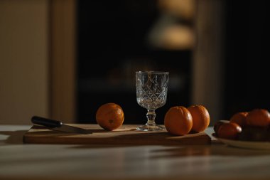 Ahşap tahta, rahat bir mutfak ortamındaki yumuşak ışıkla aydınlatılmış kristal bir camın yanında taze portakallar sergiliyor. Bıçak portakalların yanında duruyor ve atmosferi güzelleştiriyor..