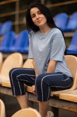 brunette girl smiles while sitting in the empty stands of a stadium. Dressed in a blue T shirt and comfortable pants she captures a peaceful moment amidst the surroundings.