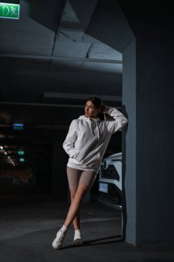girl leans against a wall in an underground parking area. She wears a white hoodie and poses casually showcasing a relaxed yet confident vibe in low light.