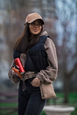 girl in a cap strolls through a park dressed in a cozy outfit. She holds a drink in one hand and appears relaxed while enjoying the fresh air.