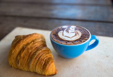 Kruvasan ile mavi Kupası'nda sıcak cappucino ahşap masa arka plan üzerinde bu