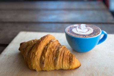 Kruvasan ile mavi Kupası'nda sıcak cappucino ahşap masa arka plan üzerinde bu