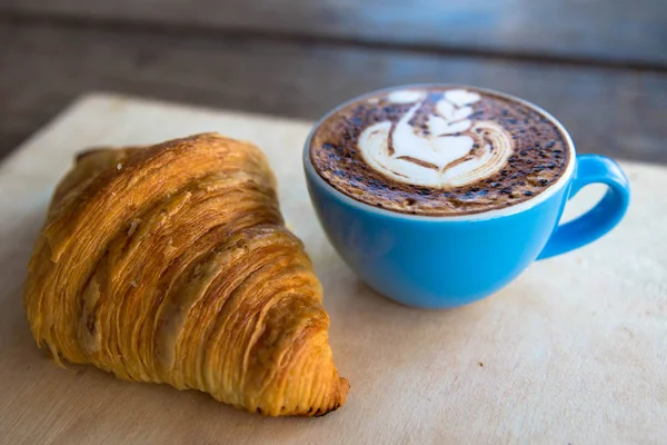 Kruvasan ile mavi Kupası'nda sıcak cappucino ahşap masa arka plan üzerinde bu