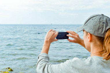 Kayalık sahilde akıllı telefonlu gri kapüşonlu genç bir kadın.