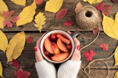 Beyaz süveterli kadınların elleri, ahşap arka planda ılık meyve suyu ve sonbahar sarısı ve kırmızı yapraklı fincanları tutarlar..