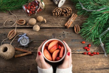 Kadınların elleri, Noel oyuncakları, böğürtlenler, tarçın ve çam dallarıyla çevrili koyu ahşap bir arka planda bir kupa şarap tutuyor..