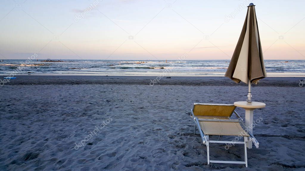 umbrellas and deckchairs closed at dawn on the rimini of rimini with sun rumbrellas and deckchairs closed at dawn on the rimini of rimini with sun reflecting off the wateeflecting off the wate. High quality photo