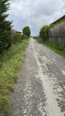 Dar ve yıpranmış bir asfalt yolu bulutlu bir günde kırsal bölgeye çıkar. Kırsal şerit, bulutlu bir gökyüzünün altında uzun yeşil çimenler, çitler ve ağaçlarla kaplı, sessiz ve huzurlu bir manzara yaratıyor. Yüksek kalite fotoğraf