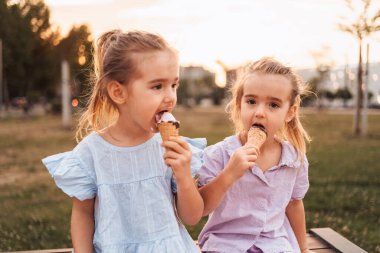 İki küçük kız bir bankta oturmuş, gün batımında bir parkta dondurma külahlarının tadını çıkarıyorlar.