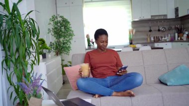 Young woman enjoying a relaxing moment on the sofa, using her smartphone while drinking coffee