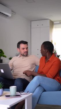 Interracial couple shopping online using laptop and credit card, comfortably seated on their sofa at home