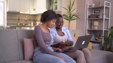 Two young women are sitting on a couch at home, using a laptop and a credit card to shop online