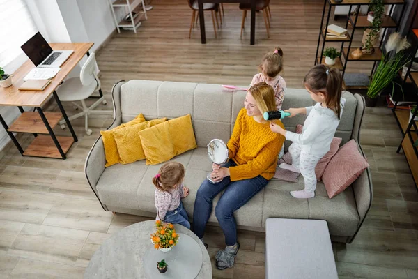 Oturma odasındaki kanepede annelerinin saçını yapan üç küçük kız gülüyor ve anneleriyle oynamaktan zevk alıyorlar..