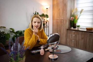 Kapalı bir alanda bitkilerle süslenmiş, sarı kazaklı bir kadın dikkatlice ruj sürüyor, makyaj malzemelerinin yayıldığı bir masaya oturuyor, tarzını yansıtıyor..