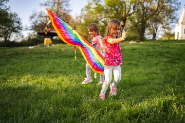 Two young girls are happily running across a lush green field, waving a vibrant rainbow flag. The sunny day brings smiles as they enjoy the beauty of nature and friendship.