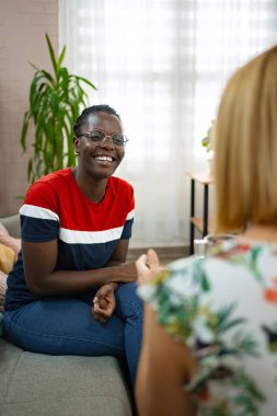 In a cozy living room filled with plants, two friends engage in a joyful conversation. One woman, wearing a colorful striped shirt, smiles warmly while listening attentively to her companion.