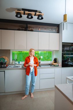 A woman stands barefoot in a modern kitchen, lost in thought.