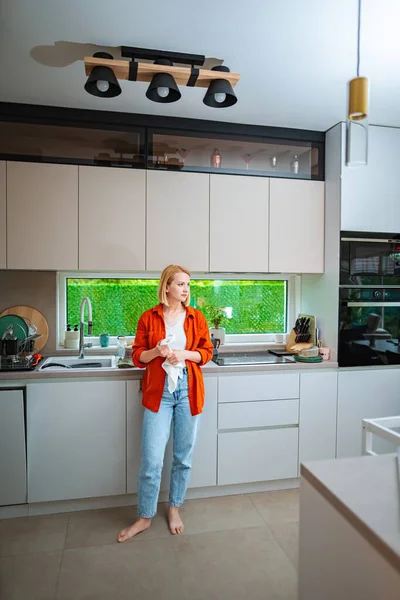 A woman stands barefoot in a modern kitchen, lost in thought.