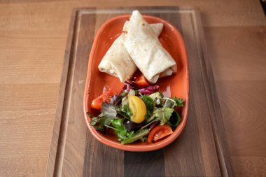 Sıcak bir yemek atmosferinde taze ve lezzeti harmanlayan canlı bir salatanın yanında, turuncu bir tabakta iki lezzetli paket dinleniyor..